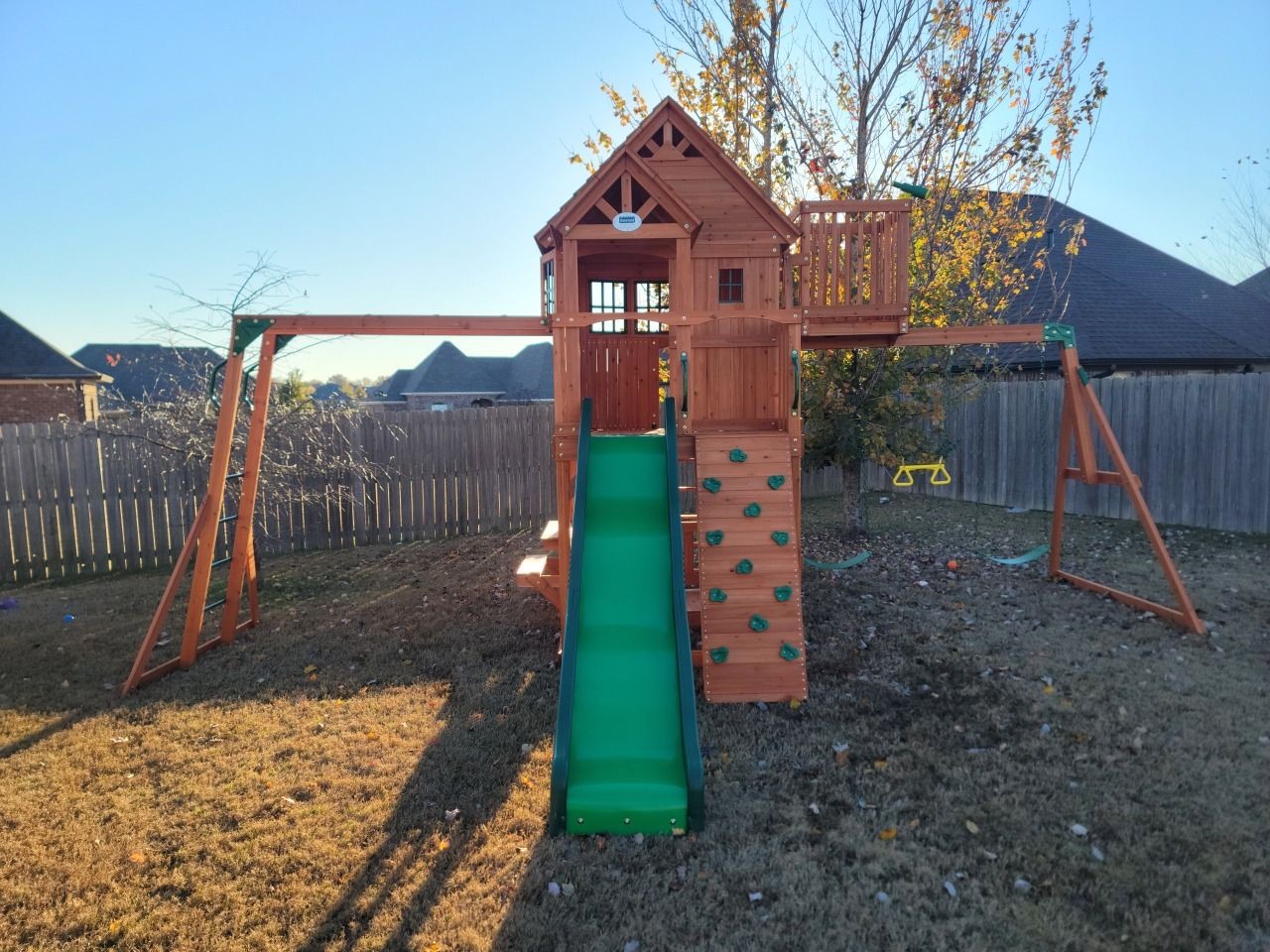 Wooden playground set with slide, swings, and climbing wall in backyard.