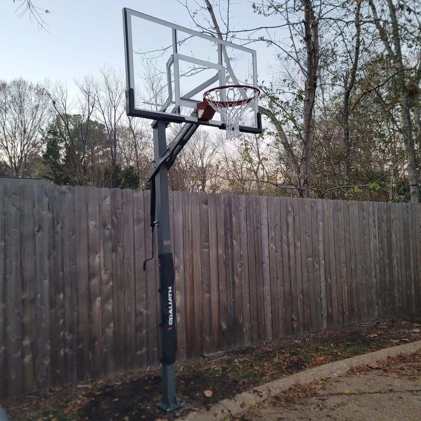 Basketball hoop with a transparent backboard near a wooden fence and trees in a backyard.