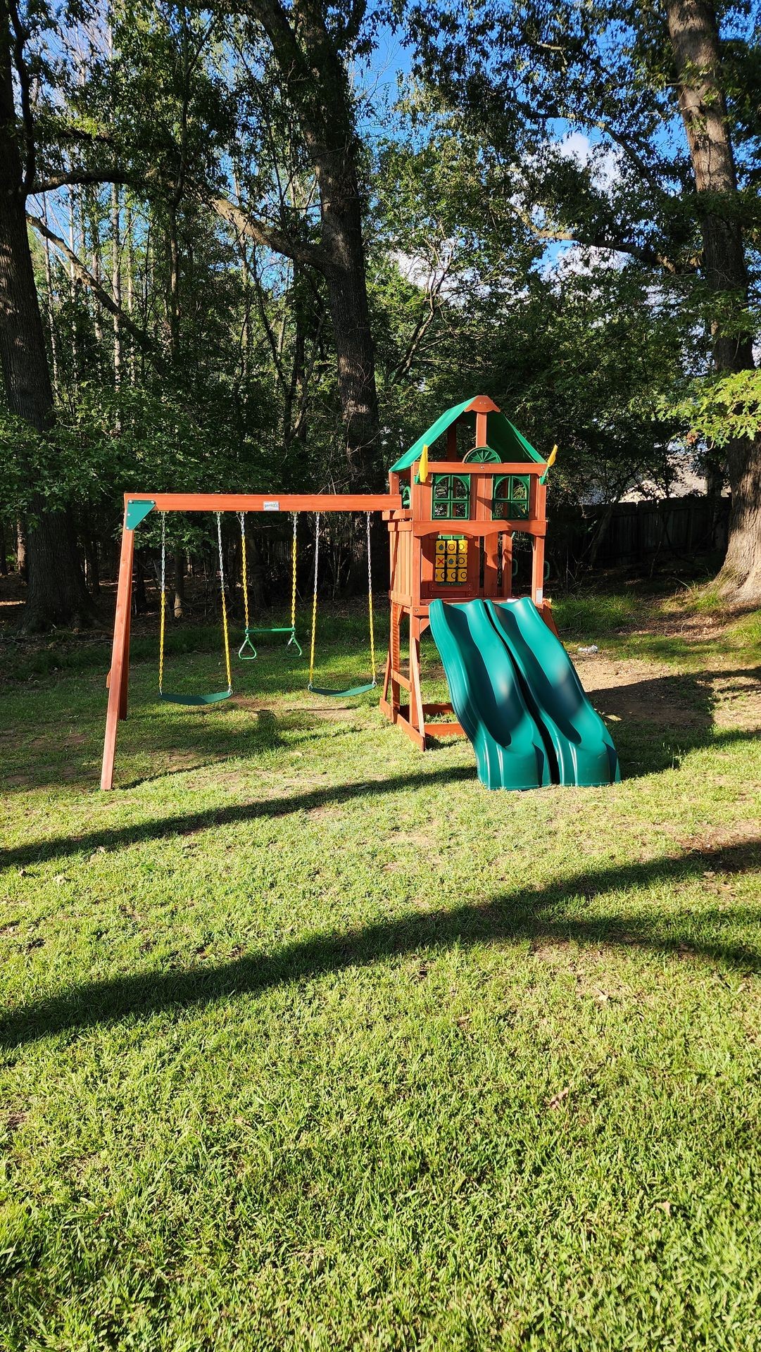 Wooden play set with swings and double slides in a sunny, grassy backyard.