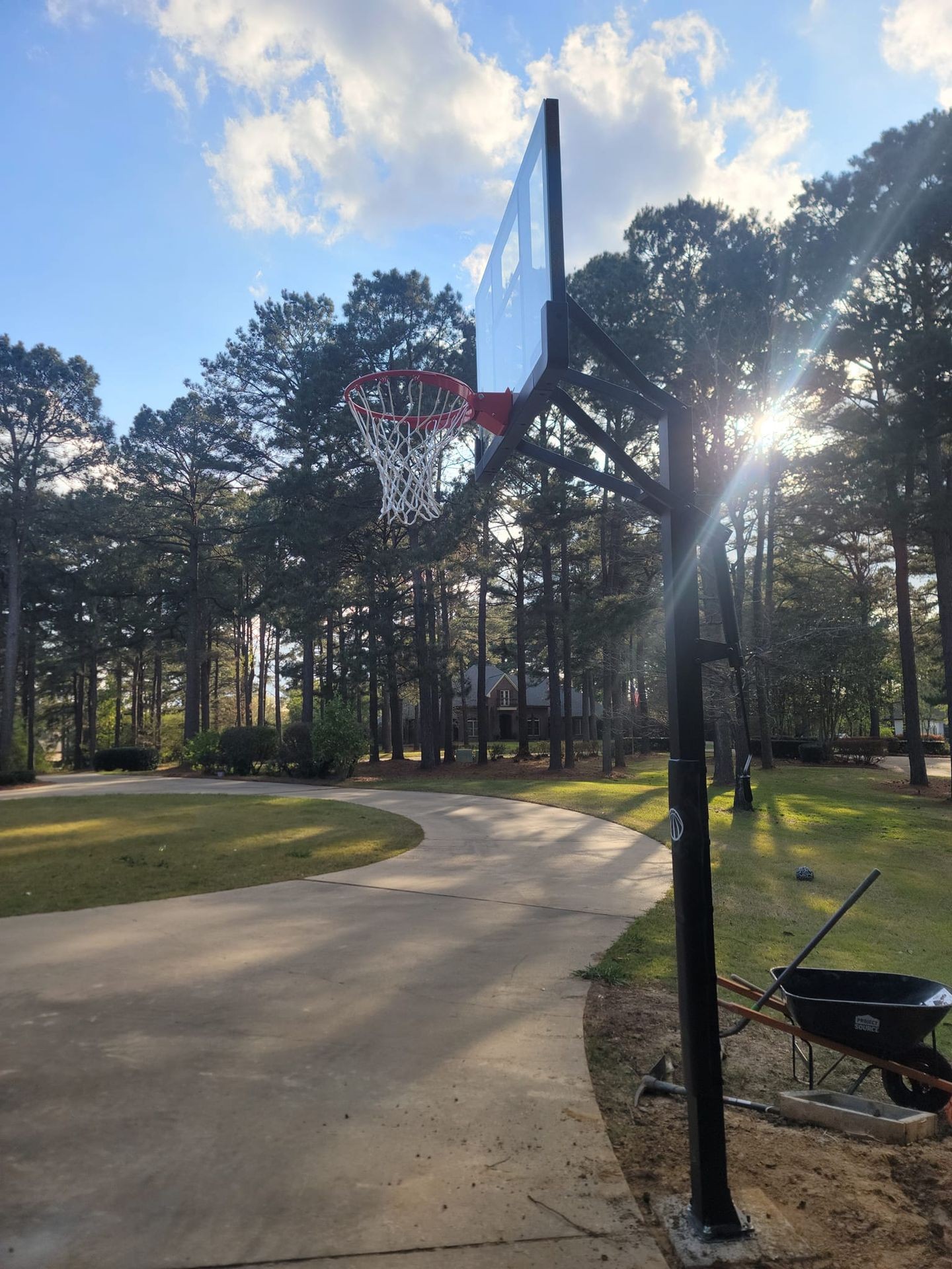 Outdoor basketball hoop with a long driveway and trees in the background under a partly cloudy sky.